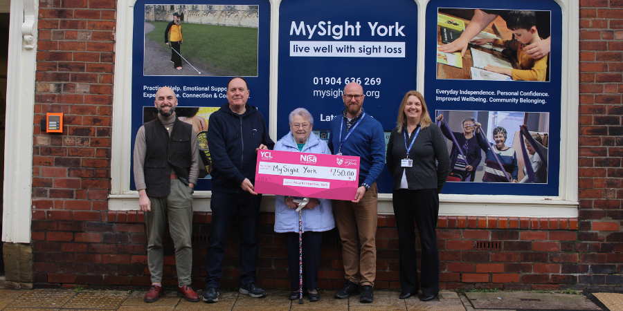 Staff and users standing outside MySight York shop holding a large pink cheque for £250. An elderly woman with a walking stick stands in the centre, smiling. The shop window behind them displays information about MySight York and support for people living with sight loss.